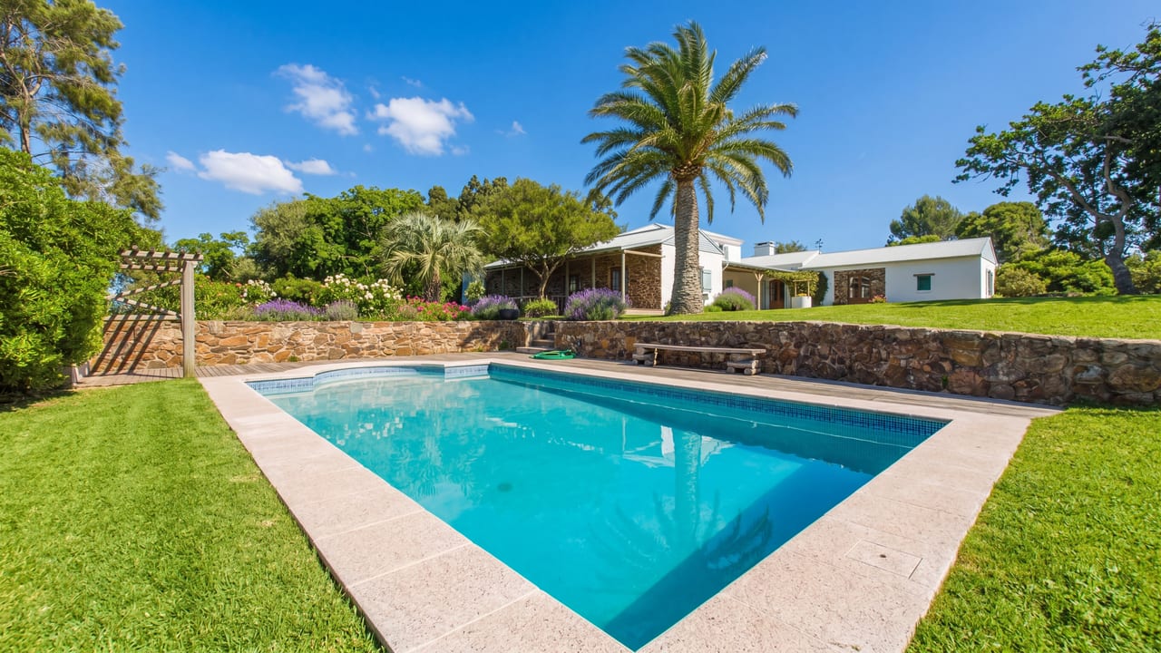 Piscina rectangular con borde de piedra y jardín con palmera, casa de campo con muros de piedra, estancia en Rocha Rural