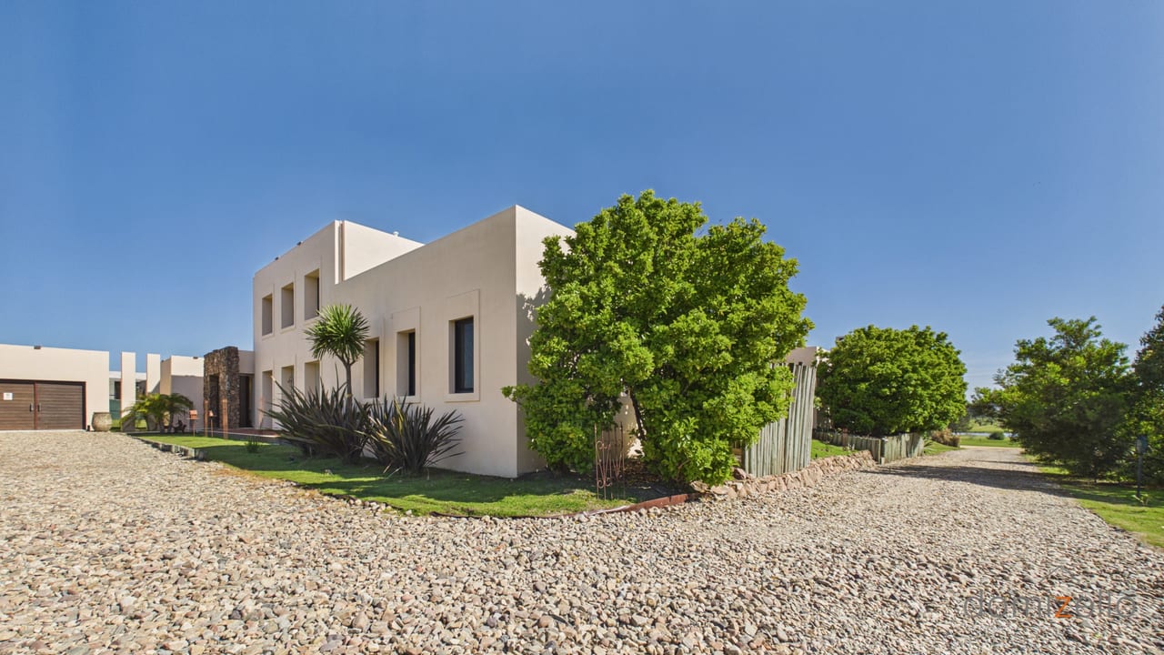 Casa moderna con fachada blanca, jardín con árboles frondosos y acceso de grava, en El Quijote, La Barra